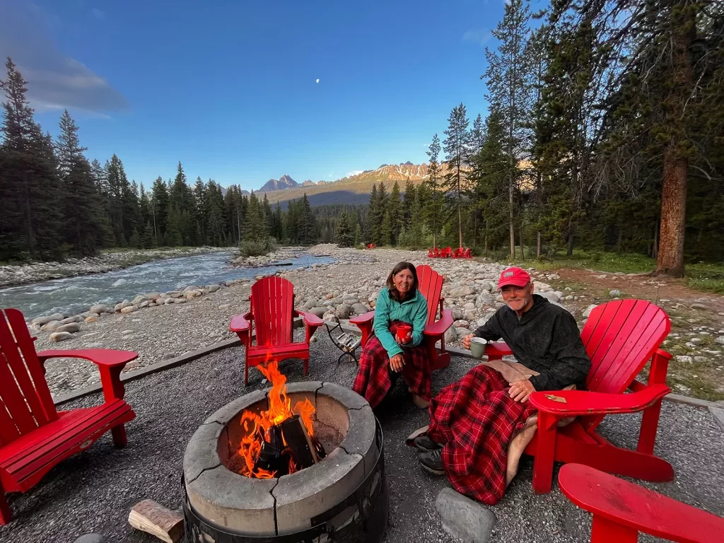 Two guests in red chairs sitting around fire, river, trees, mountain background.
