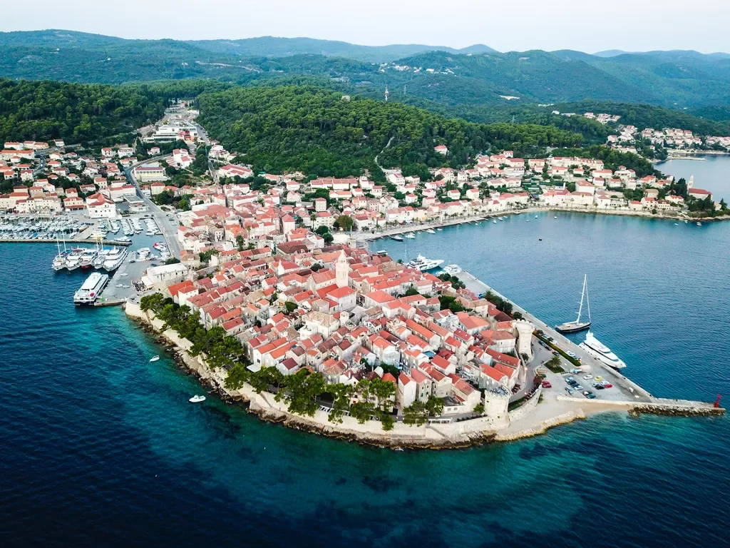 Wide shot of coastal Croatian town, white and tan buildings, boats, forest in distance. 