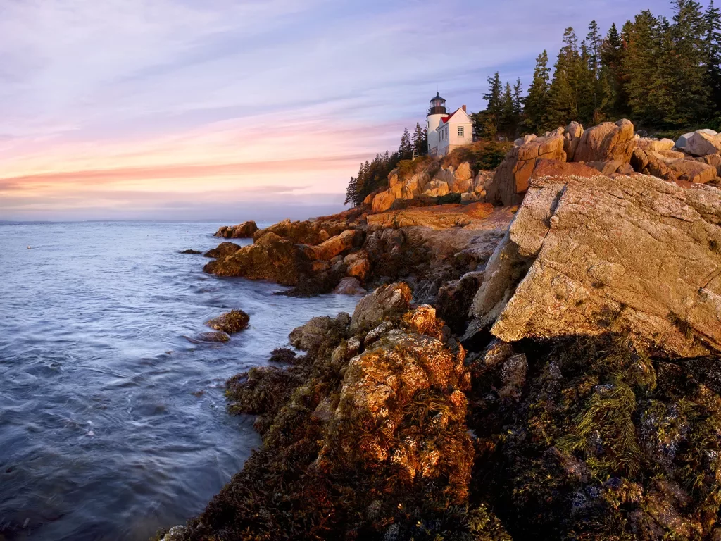 Wide shot of craggy inlet, small white lighthouse, sunset.