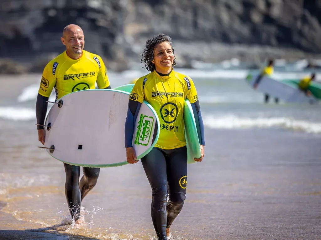 Two people in yellow rash guards carrying surfboards along the beach