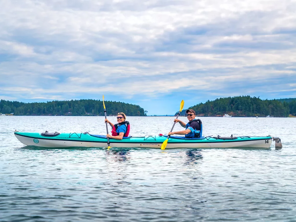 Two guests kayaking, smiling at camera, hills, sky behind them.