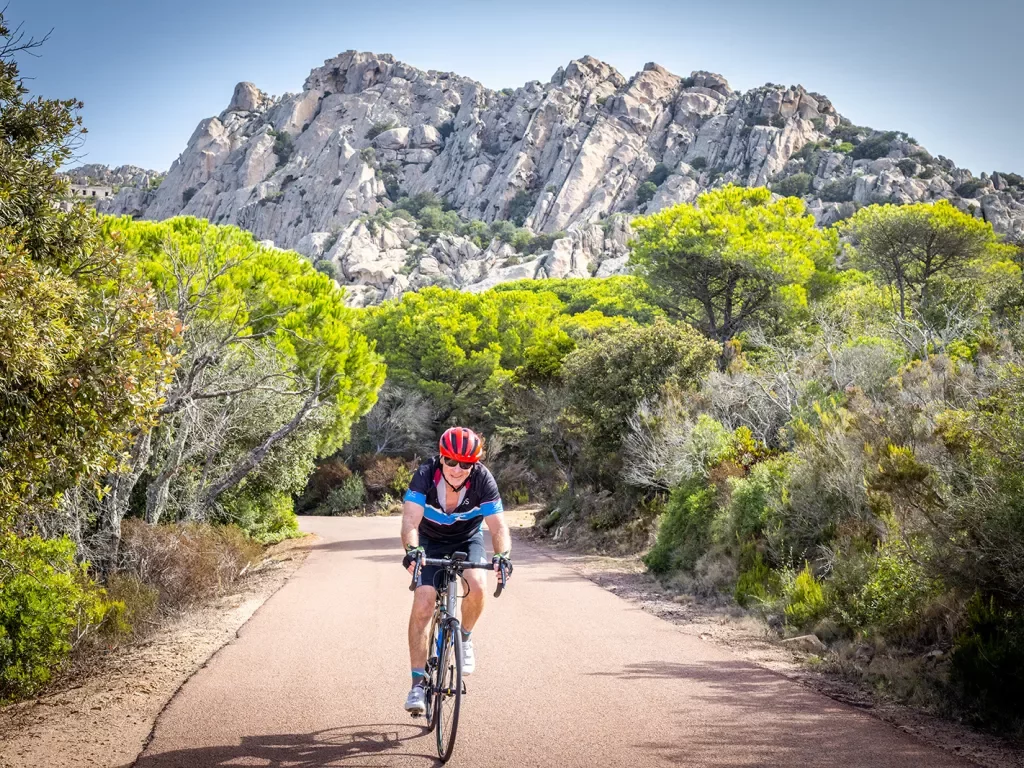 Guest cycling towards camera, trees surrounding them, rocky cliff in background.