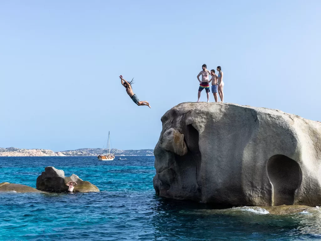 Four guests jumping off large rock, one in the water below.