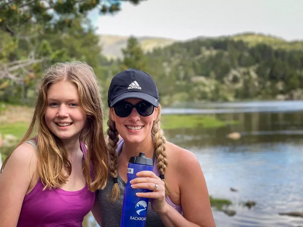 Two women smiling at the camera while hiking beside a lake