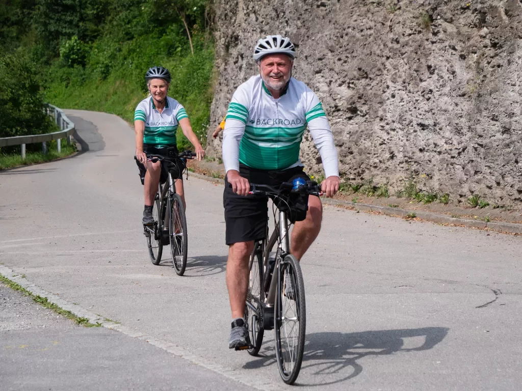 Two guests biking down road, Backroads jerseys on, smiling.