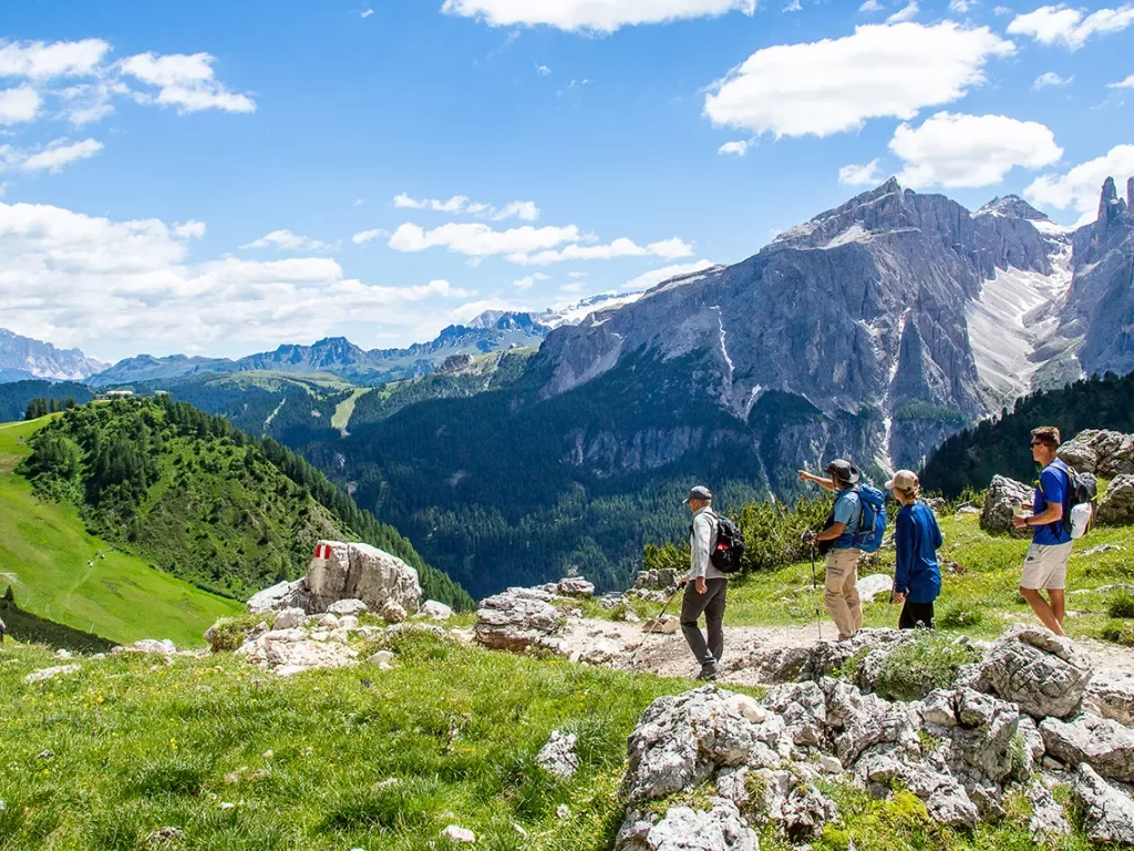 Group of guests hiking down mountain trail, Dolomite range in background.