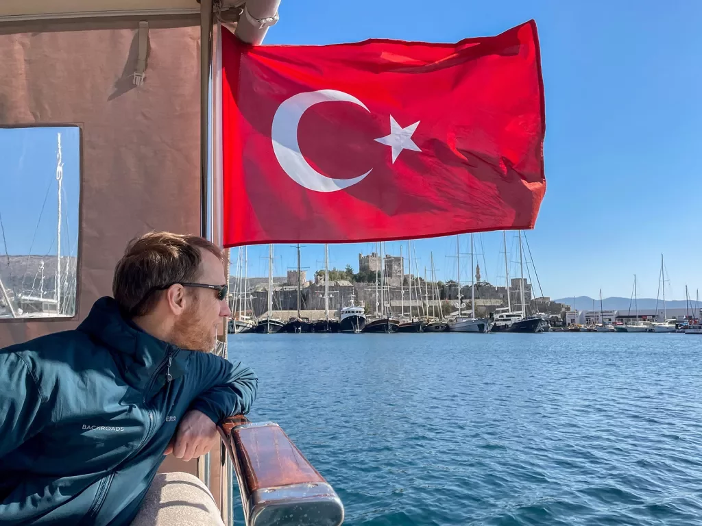 Guest on pier overlooking fleet of sailboats, Turkish flag behind him.