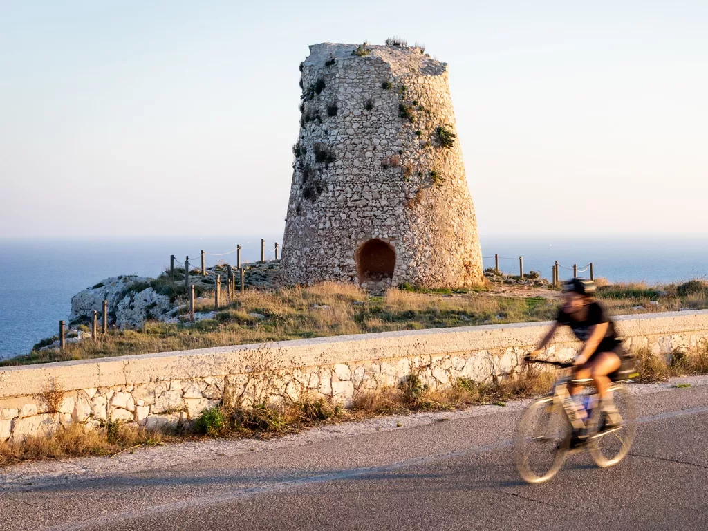Shot of guest cycling past old, crumbling tower, ocean to their right.