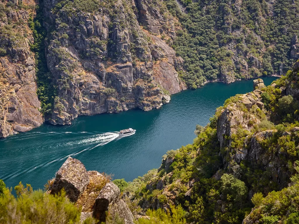 Hillside shot of craggy river valley, passenger boat in water below.