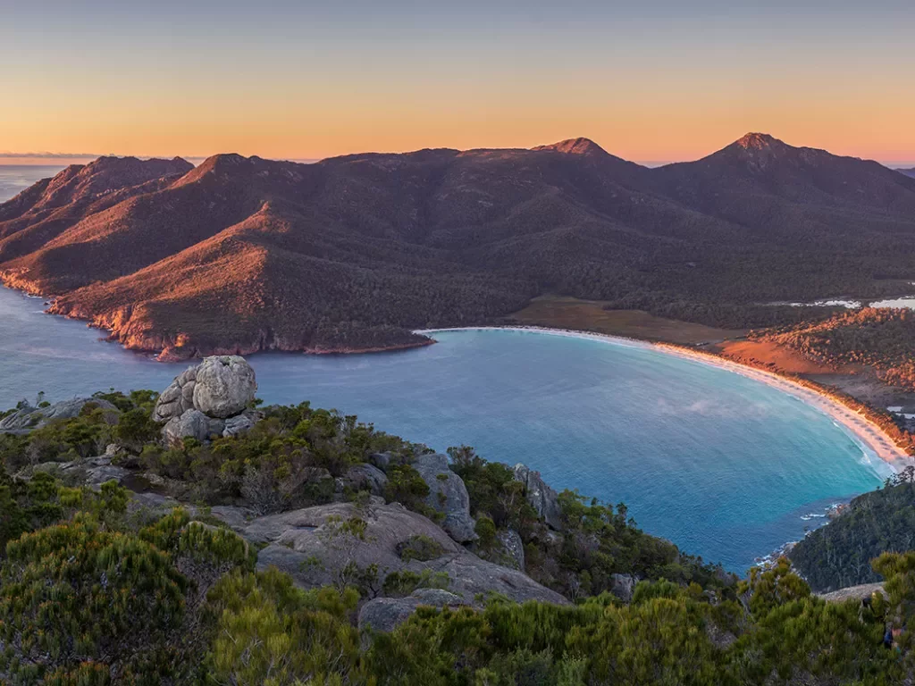 A panoramic landscape image of sunrise at the peak of Mount Amos with Wineglass Bay at the background, Freycinet National Park, Coles Bay, Tasmania, Australia.