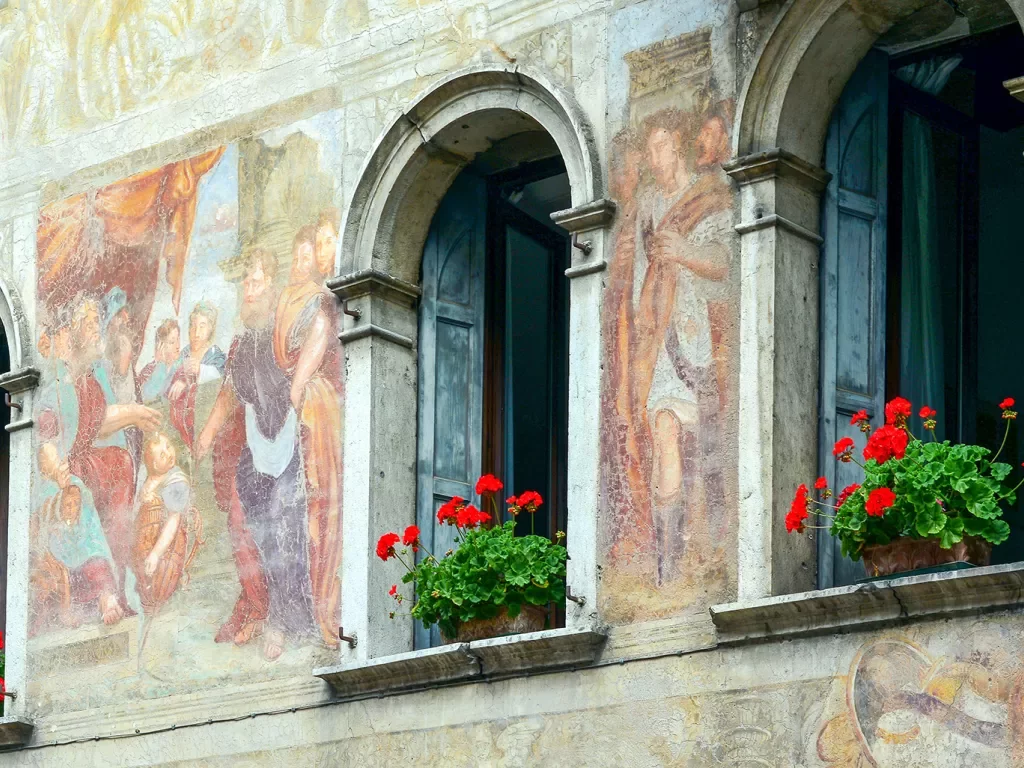 Shot of ancient mural on side of building, open windows, flowers.