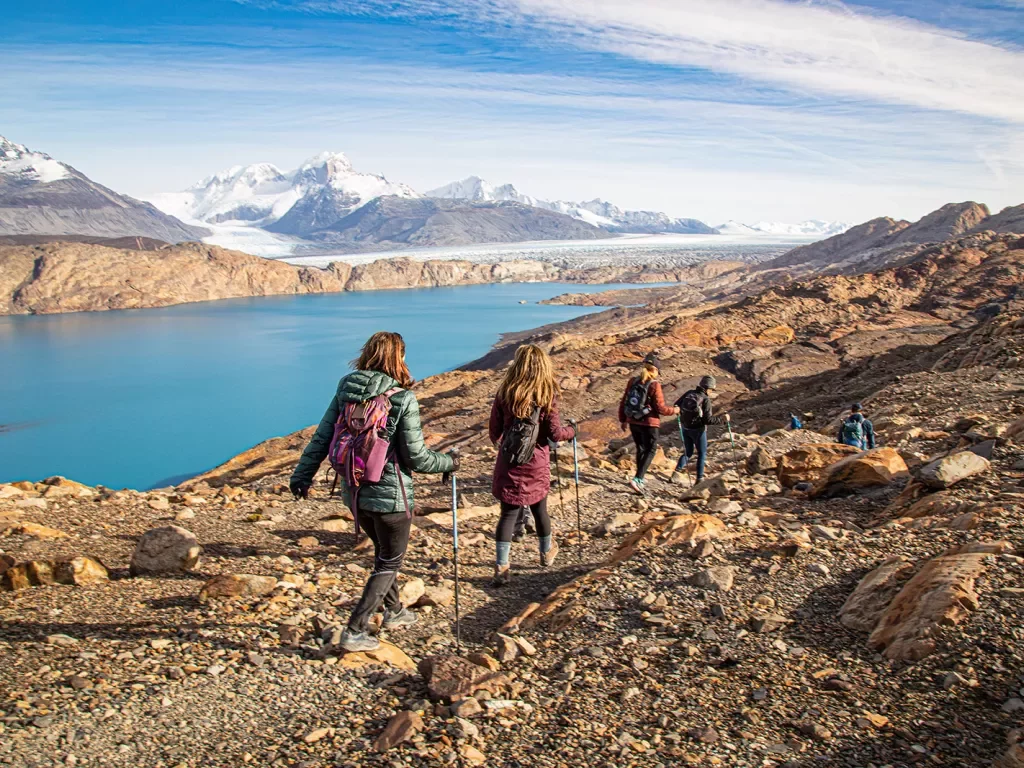 Group of guests walking towards blue lake, snowy mountains in distance. 