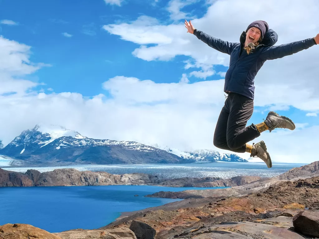 Guest on cliffside, jumping for joy. Lake, snowy mountains behind her.