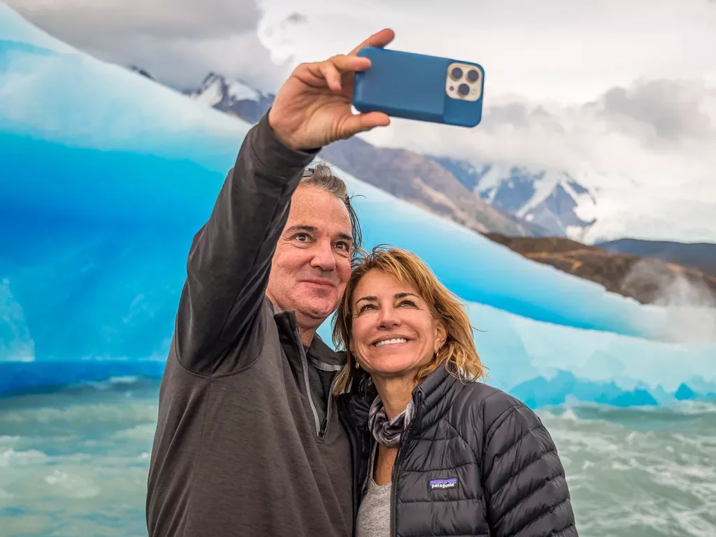 Two guests taking selfie in front of large glacier.