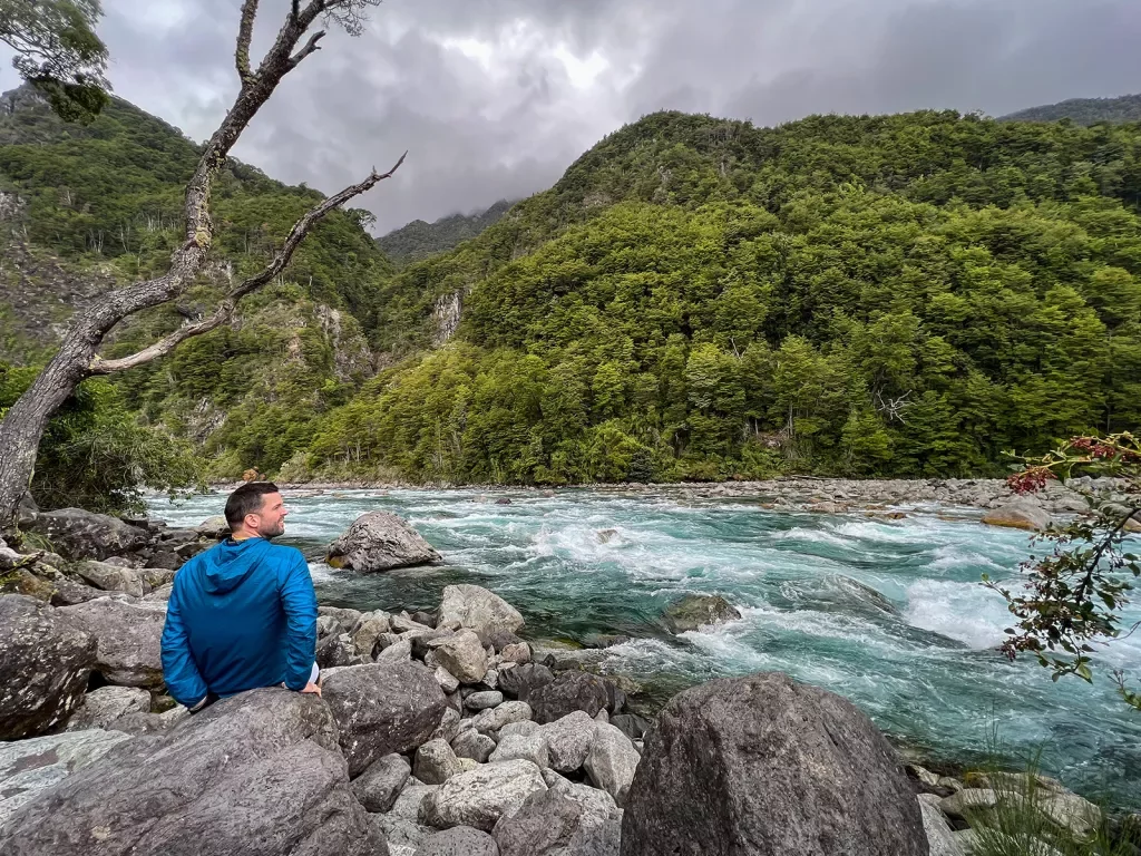 Guest sitting on riverside, white water flowing, mountains and grey clouds above.