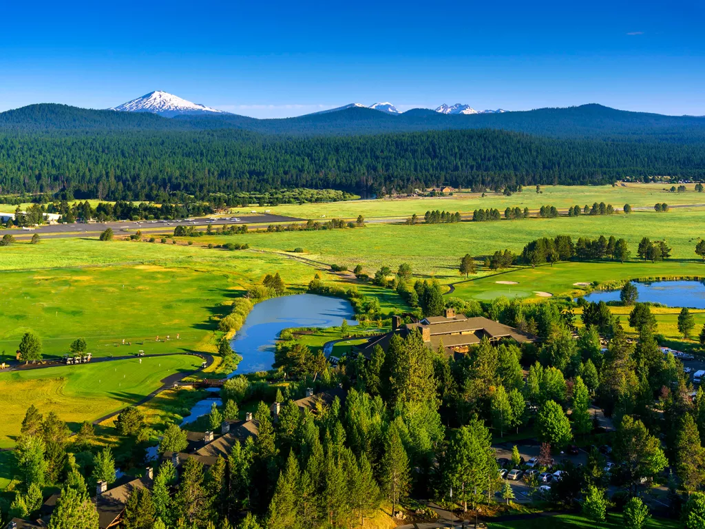 Wide shot of green pasture, large house in foreground, mountains in background.