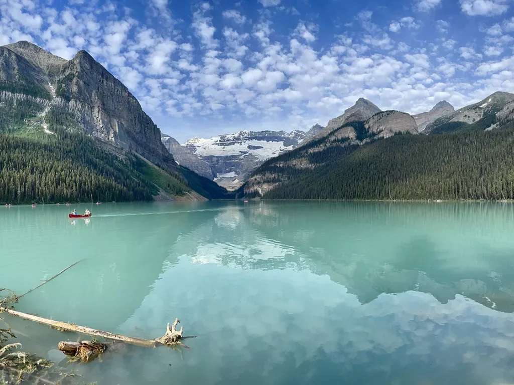 Wide shot of large blue lake, people canoeing in distance, mountains, clouds, sky in background.