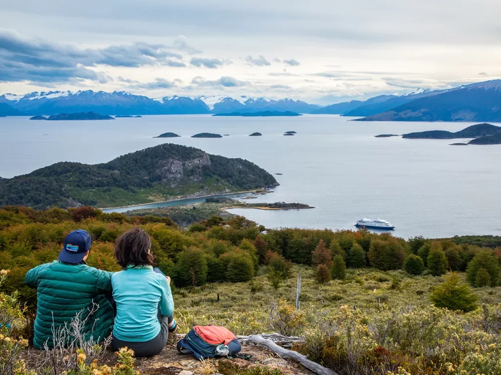 Two guests sitting on hilltop, overlooking huge lake, ring of mountains, clouds, etc.