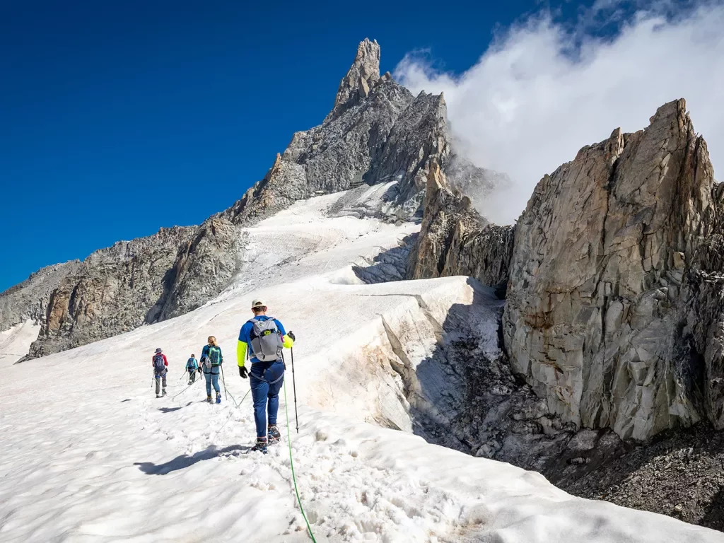 Four guests walking beside snowy cliff, sharp, craggy mountain behind them.
