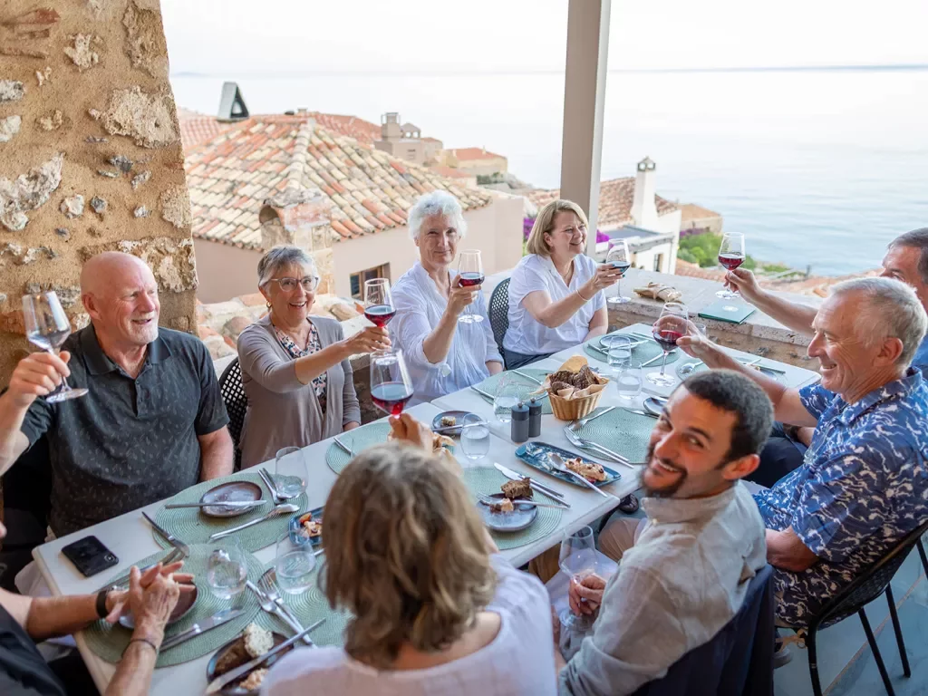 Guests at dinner table, all cheersing wine glasses, overlooking ocean.