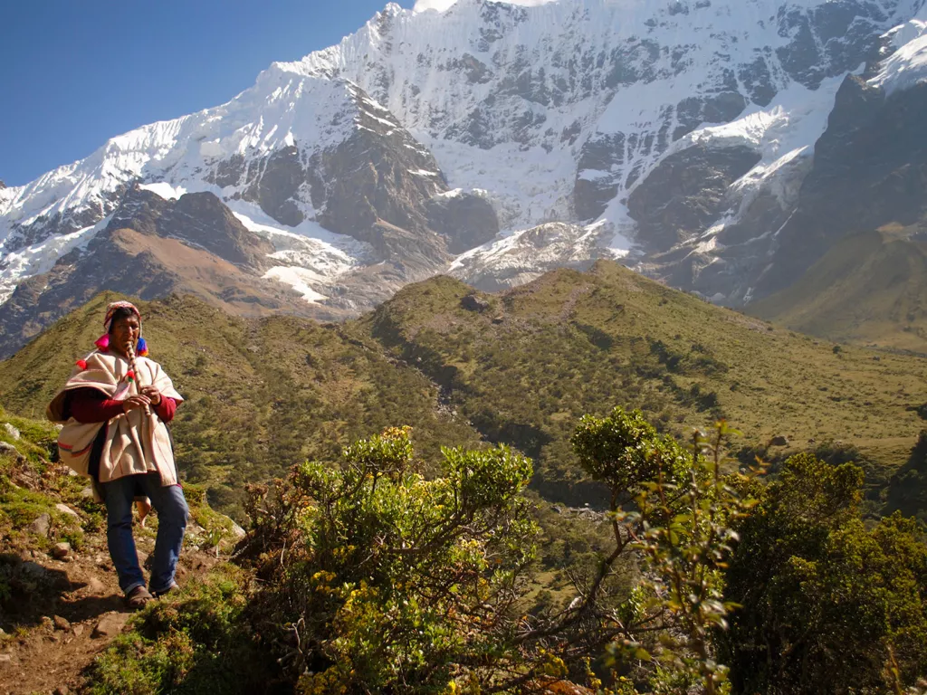 Local on hillside playing instrument, snowy mountains, hills behind them.