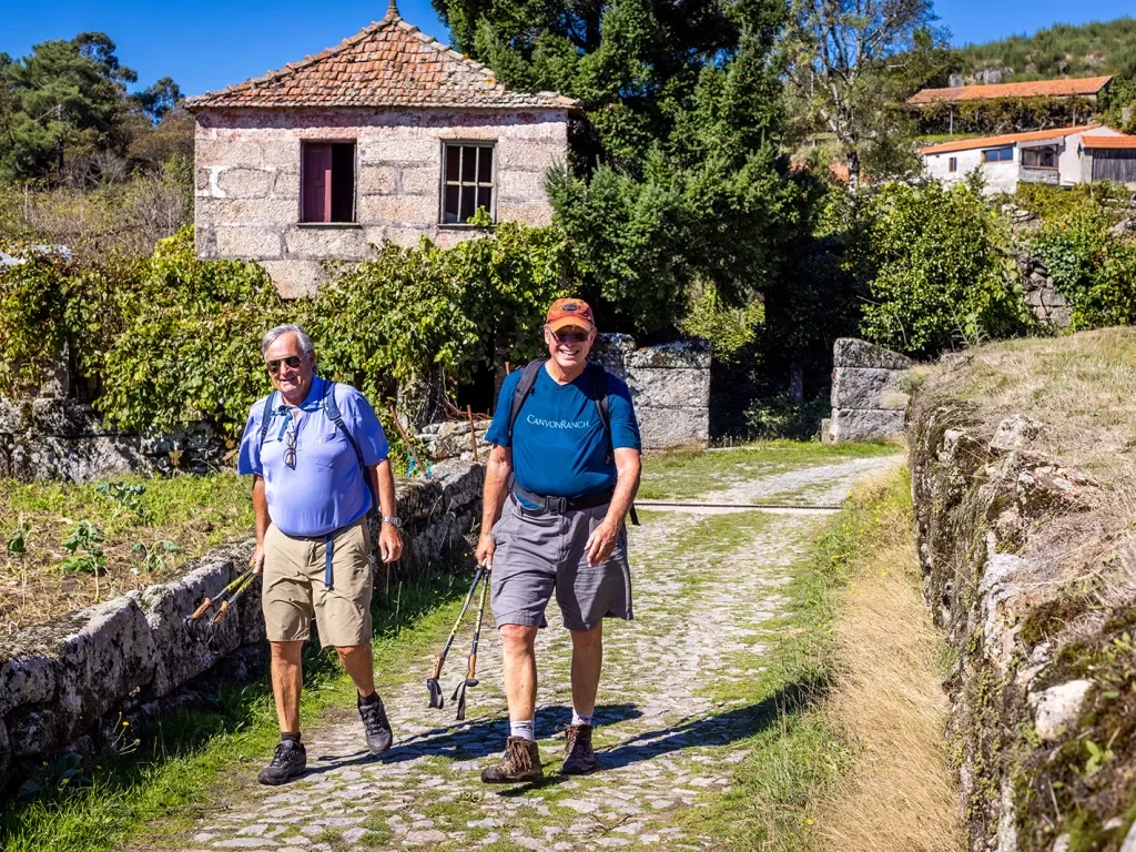 Two guests walking through countryside vineyard, stone buildings behind.