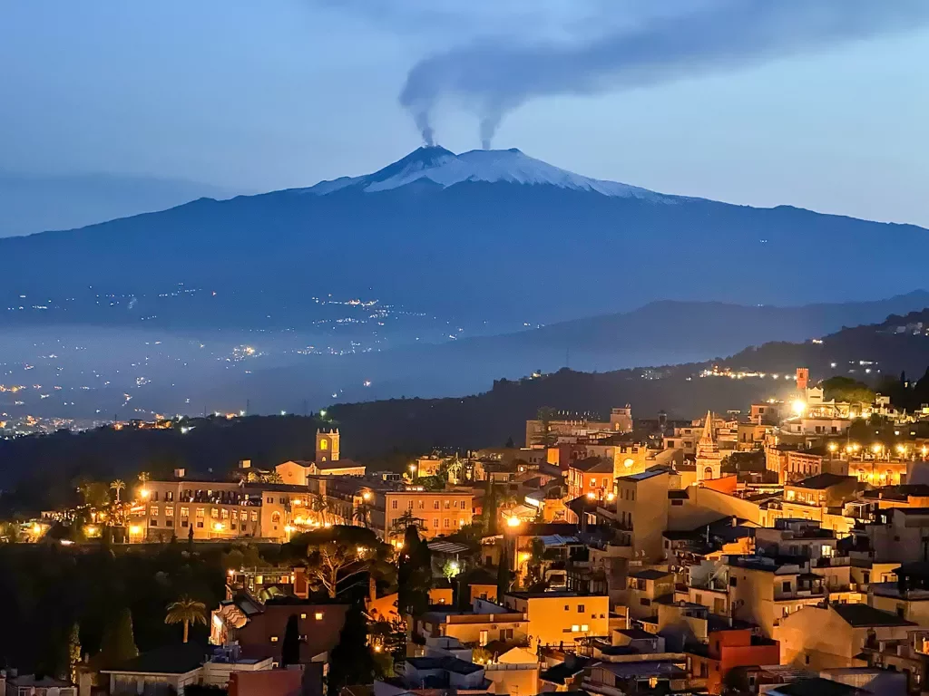 Nighttime shot of Italian hillside town, smoking volcanoes in distance.