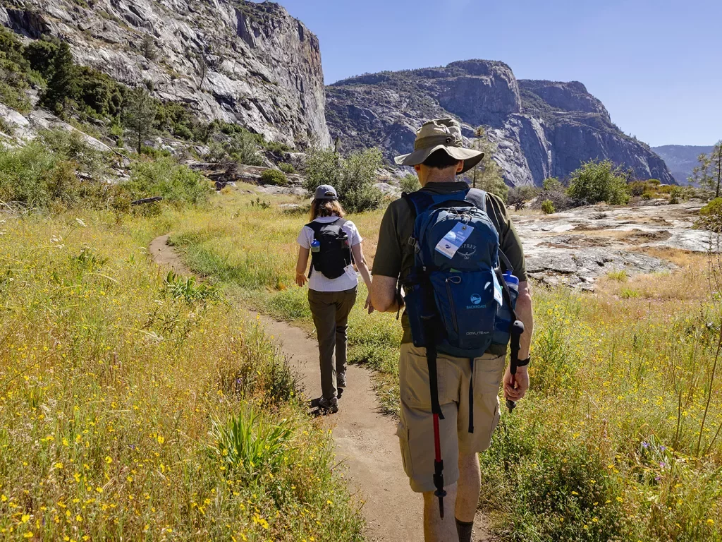 Two guests hiking on grassy trail, large cliffs to their left.