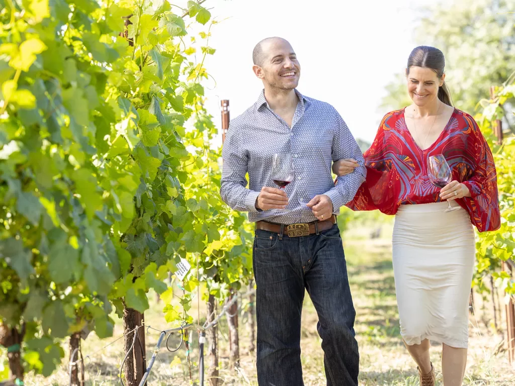 Couple walking through a vineyard, holding glasses of wine