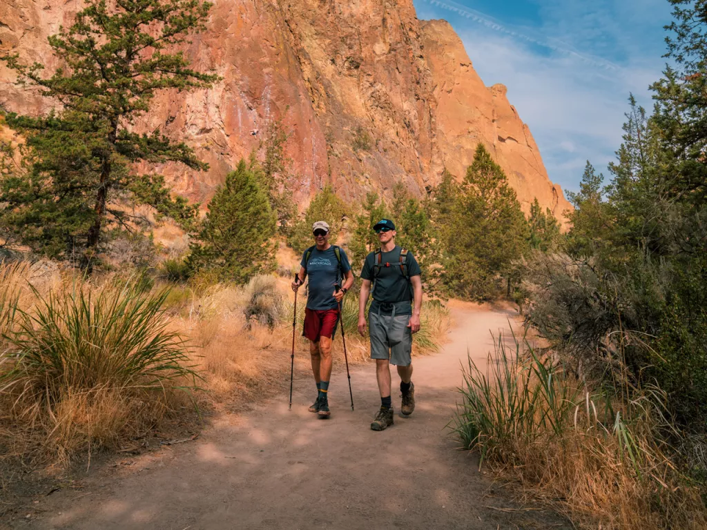 Two men with walking poles, hiking on a dirt trail next to a canyon