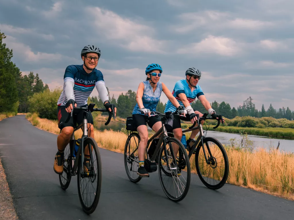 Two men and one woman riding bicycles on an asphalt road, next to a river