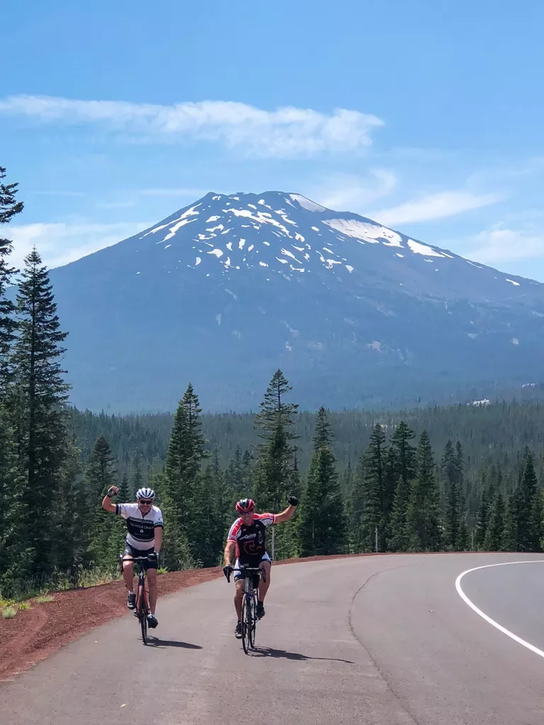 Two guests cycling and waving to camera, Mount Bachelor in background.