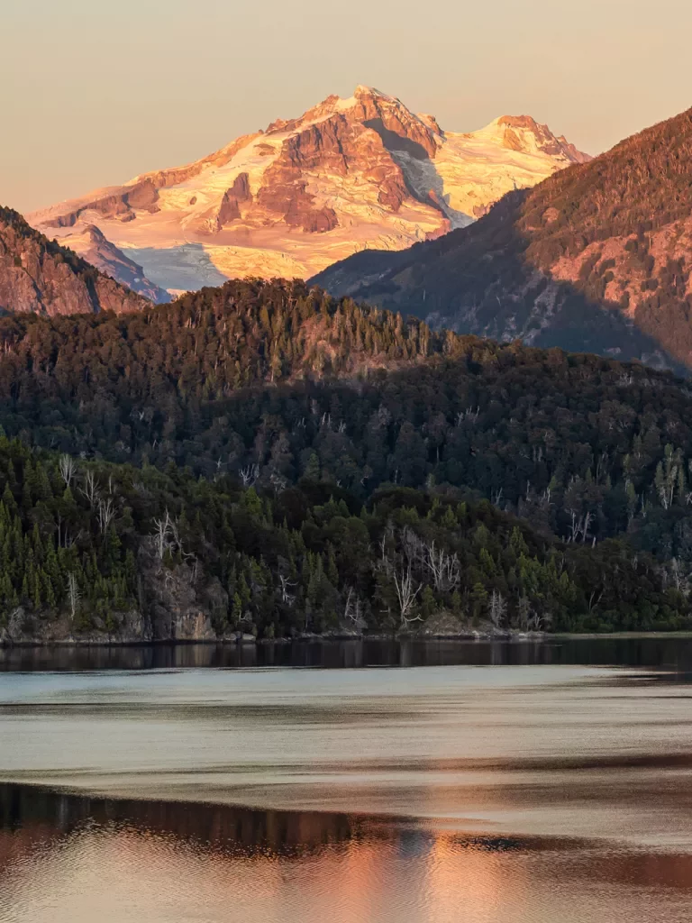 Wide shot of reflective lake during sunset, forest, snowy mountain behind.