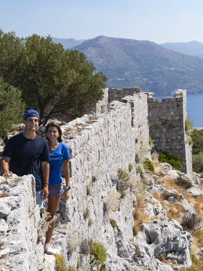 Two guests on ruins of white-rock wall, ocean to their left.