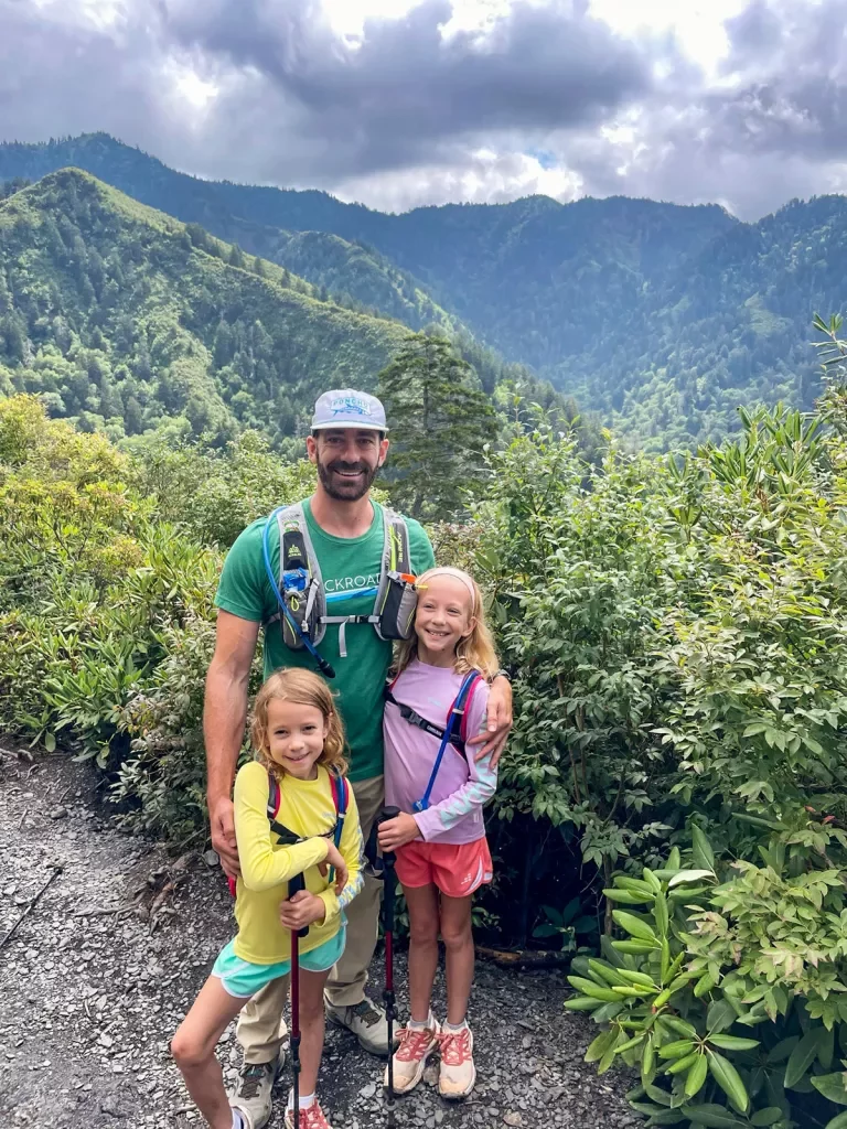 Father and two daughters smiling for camera, large forest and mountain in background.