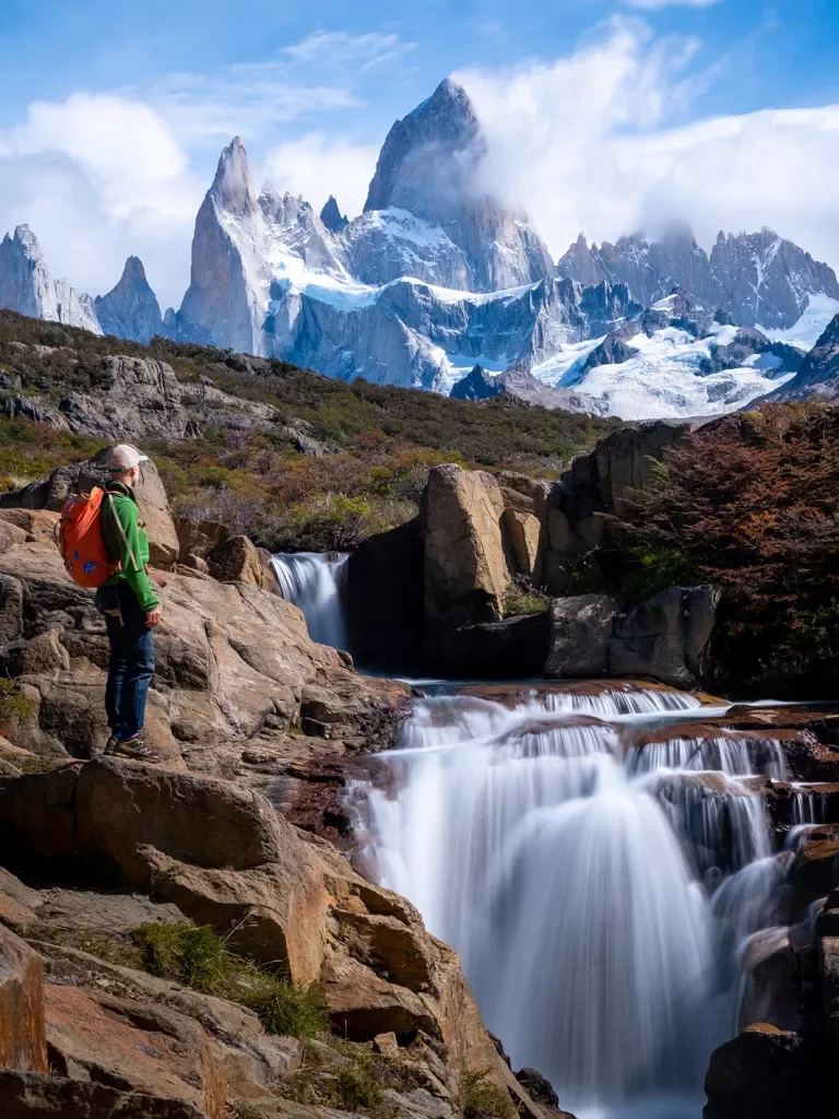 Guest standing next to flowing waterfall, sharp, snowy peaks in background.