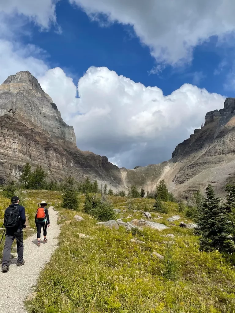 Two guests walking down meadow trail, two large mountain faces in distance.
