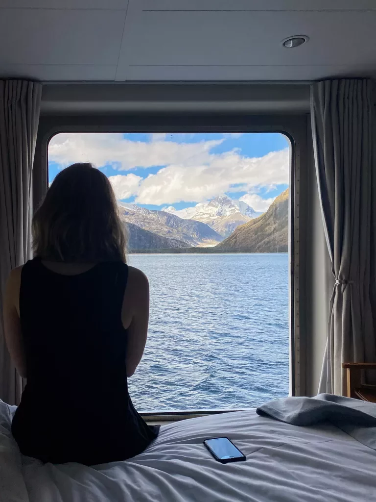 Guest on cruise ship in room, looking out towards mountains.