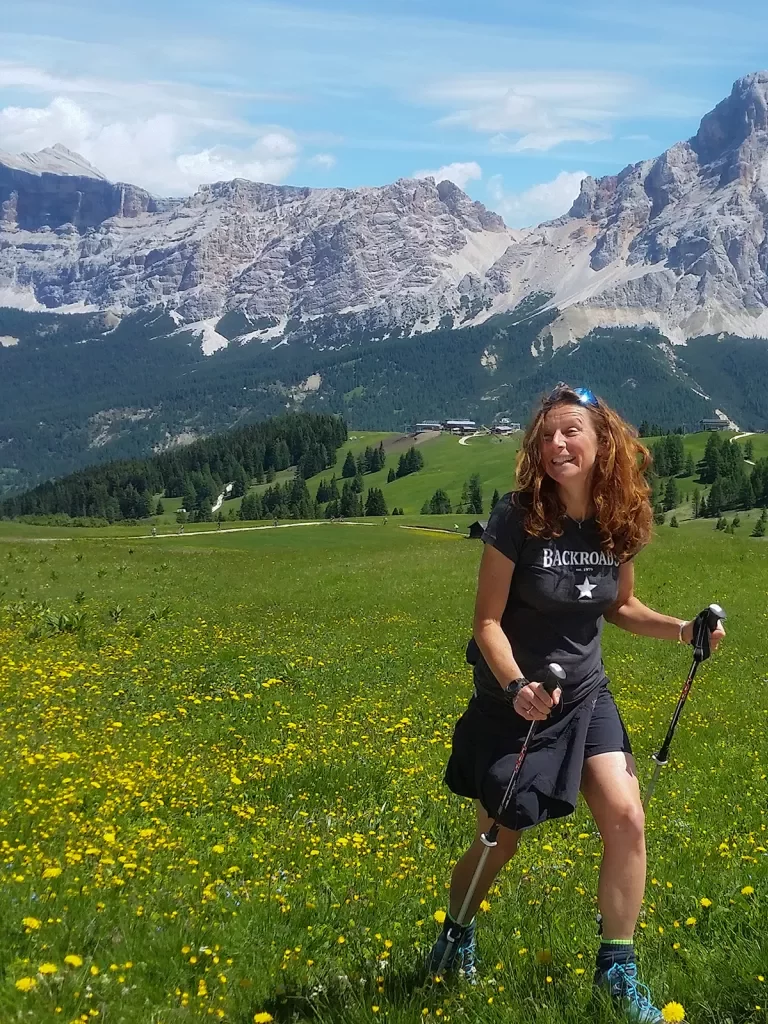 Guest with hiking poles in grassy meadow, large, grey cliffs behind her.