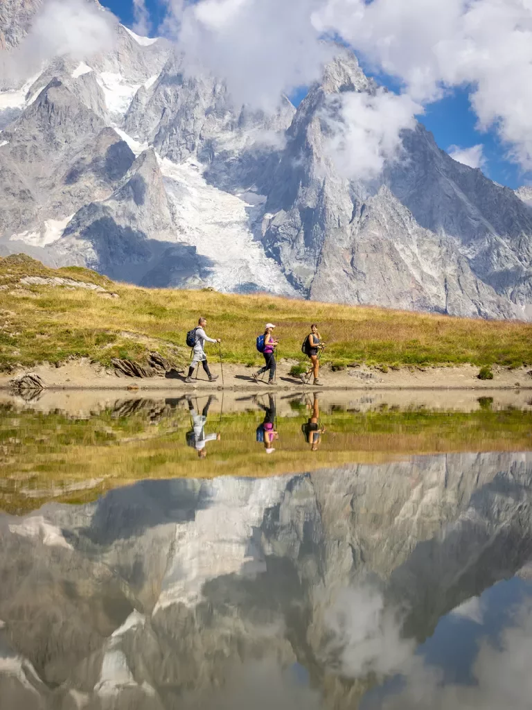 Three guests walking past reflective lake, meadow, snowy mountain in distance. 