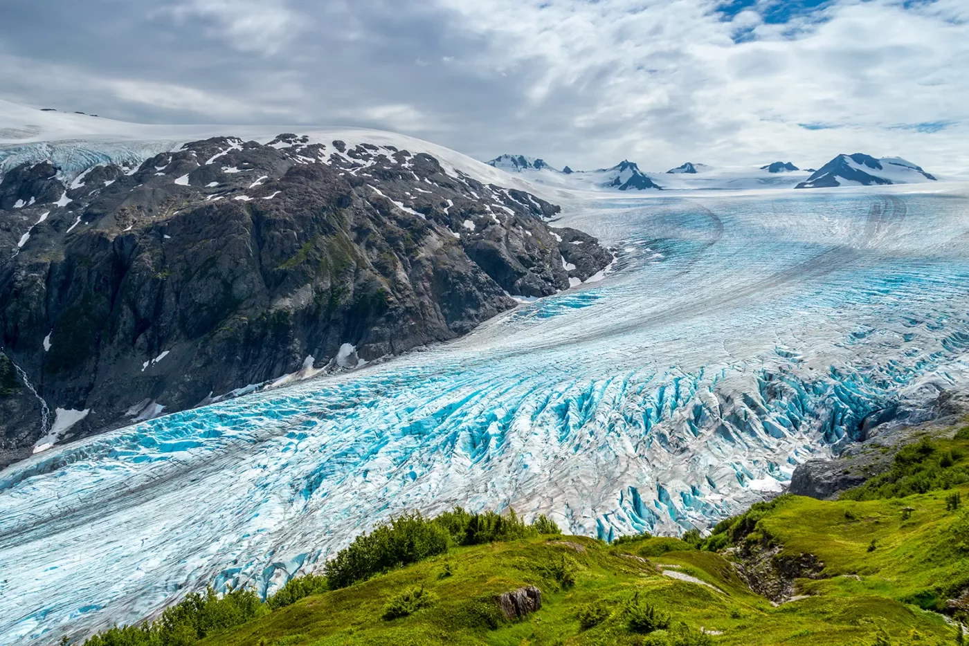 Shot of glacial vista, green cliff, blue ice, grey mountain.