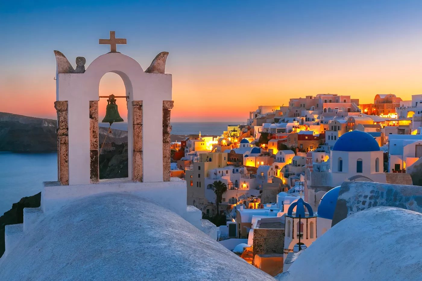 Sunset shot of Santorini, city lit up, church bell in foreground.